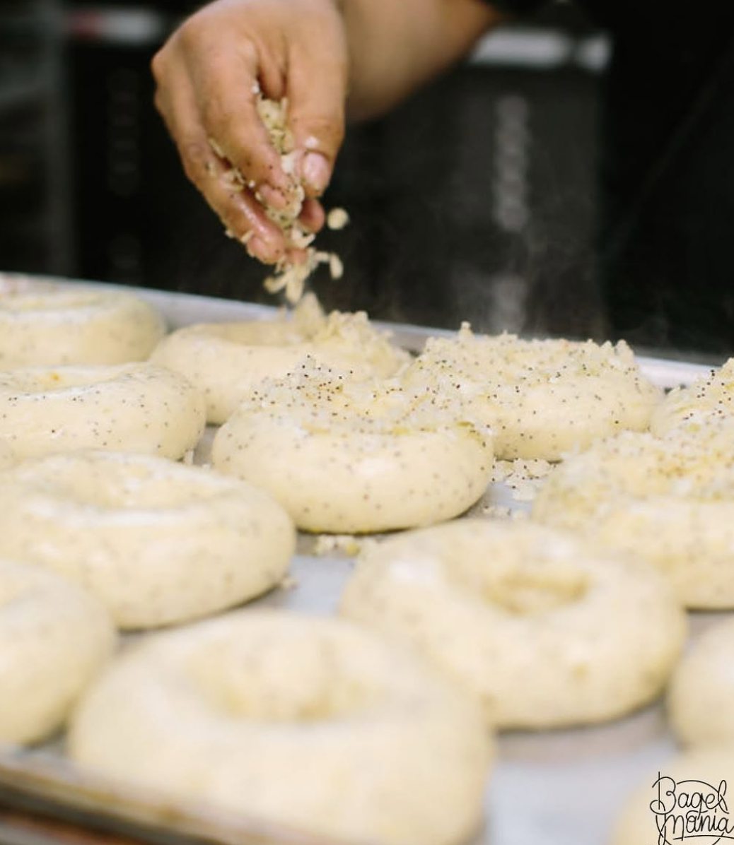 A baker sprinkling seasoning over a fresh tray of hand-rolled bagels